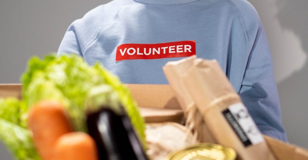 Smiling woman holding a box filled with various vegetables and canned goods during a volunteer drive.